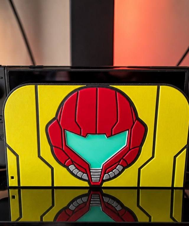 Multicolour 3D-printed Switch 2 dock faceplate with a Metroid-inspired helmet design, reflected on a glass shelf